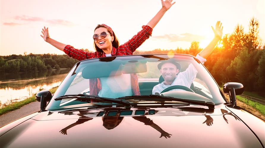 Excited couple riding in a convertible with the top down and their arms raised headed out for a long-weekend vacation. 