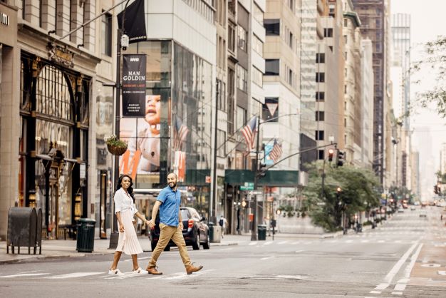 A couple holds hands and crosses the street in New York City, skyscrapers visible behind them