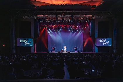 A group of people watch a signer and two guitarists perform on stage at an HGV Live! event