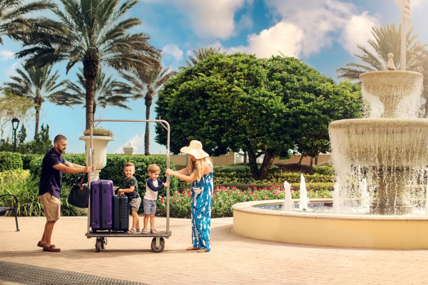 Family of four using a trolley to take their luggage across a plaza with a fountain